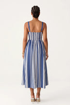 Woman wearing an A-line midi dress features a bold, vertical stripe pattern in alternating royal blue and white that creates a slimming and elongating visual effect. Designed with a structured square neckline, thick supportive straps, and a solid blue upper border, the fitted bodice gracefully transitions into a flared, flowing skirt that falls just below the calves.