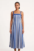 Woman wearing an A-line midi dress features a bold, vertical stripe pattern in alternating royal blue and white that creates a slimming and elongating visual effect. Designed with a structured square neckline, thick supportive straps, and a solid blue upper border, the fitted bodice gracefully transitions into a flared, flowing skirt that falls just below the calves.