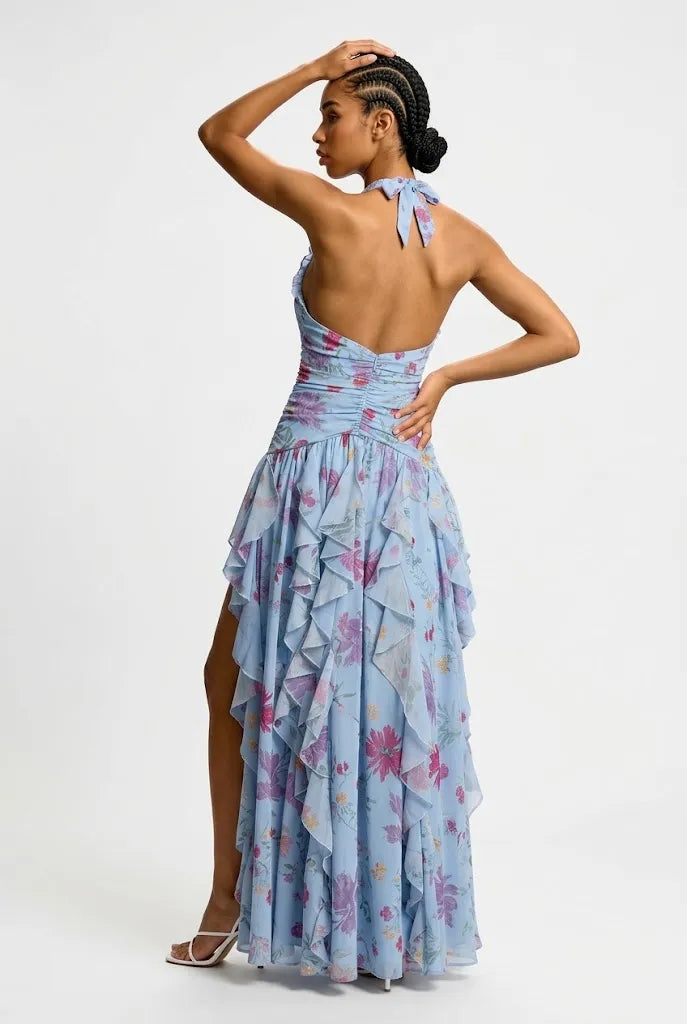 A model wearing a light blue halter-neck maxi dress with a pink and purple floral print, featuring a ruffled tiered skirt and a high side slit, set against a plain light gray background.