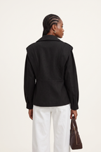 Person wearing a black jacket and white pants, holding a brown bag on a light background