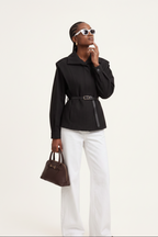 Woman wearing a black coat and white pants, holding a brown handbag on a light background
