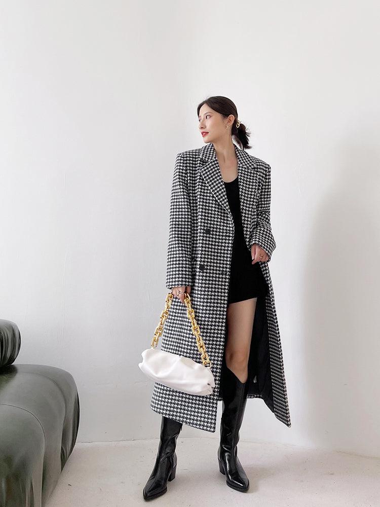 Woman wearing a long houndstooth coat holding a white handbag against a plain background