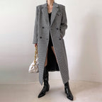 Person wearing a long houndstooth coat with black boots and a white bag against a plain background