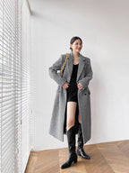 Woman in a checkered coat and black knee-high boots standing in a room with wooden flooring and white walls.