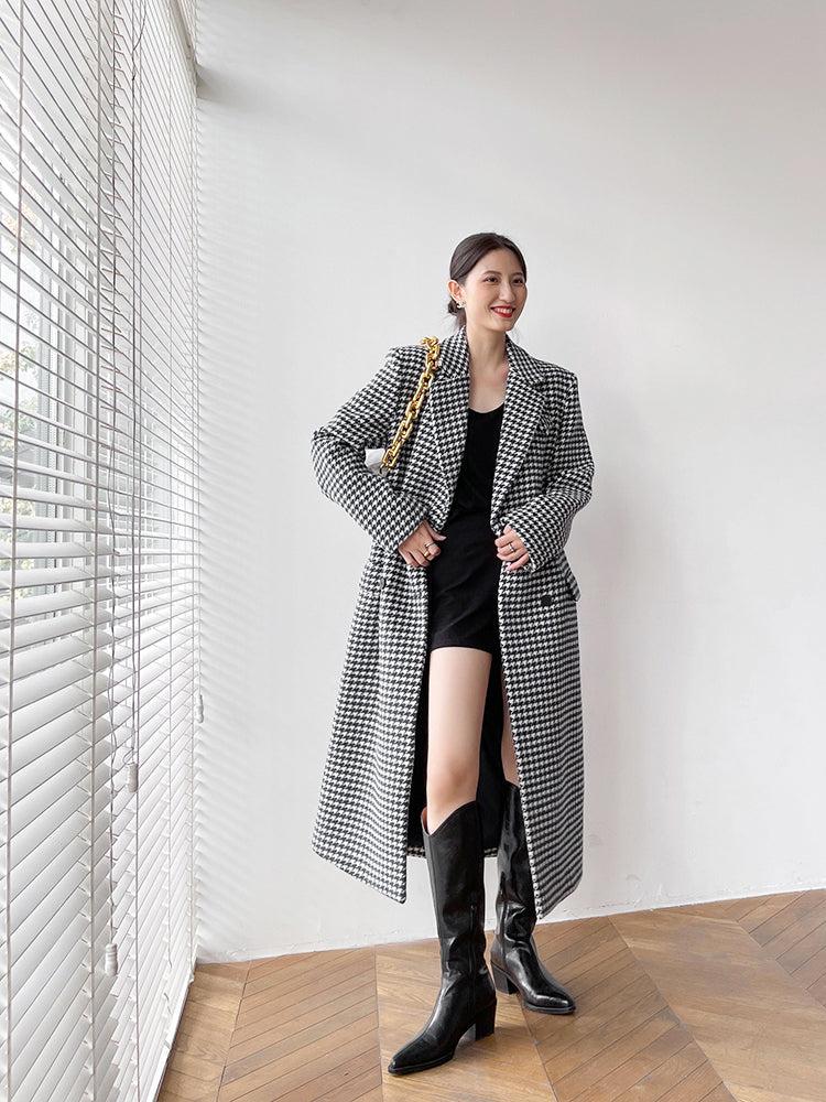 Woman in a checkered coat and black knee-high boots standing in a room with wooden flooring and white walls.