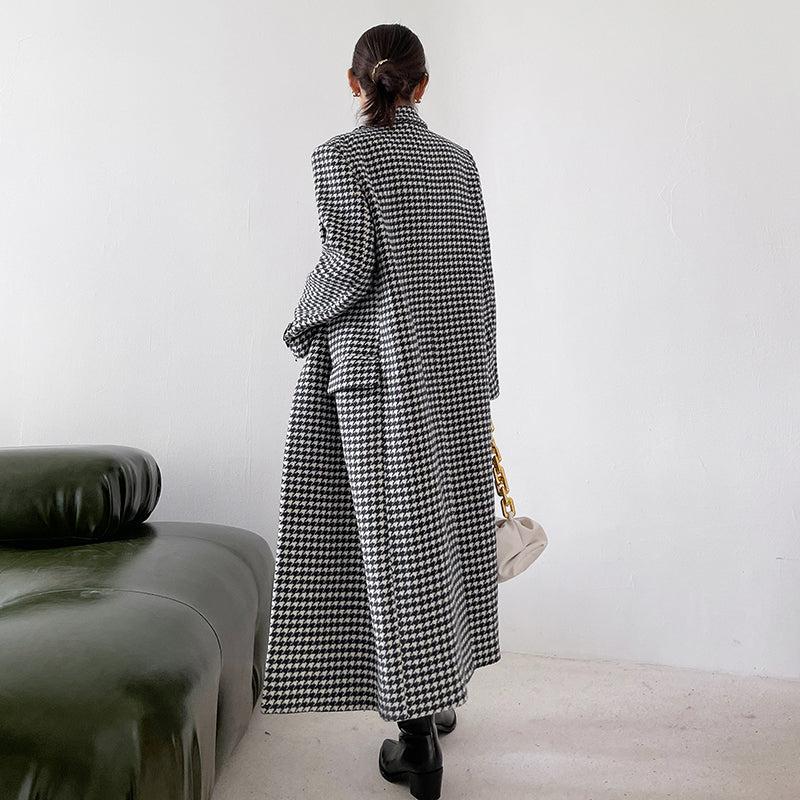 Person wearing a long black and white checkered coat standing in a minimalistic room.
