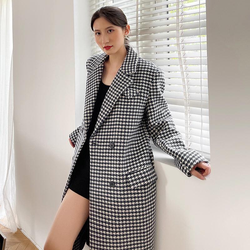 Woman wearing a black and white checkered coat standing indoors with light streaming through blinds.