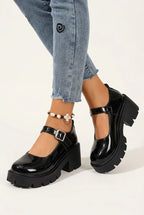 A person wearing black patent leather Mary Jane platform shoes with chunky lug soles and buckle straps, paired with light blue cropped jeans on a clean white studio background.