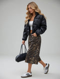 Woman wearing a black puffer jacket, white top, leopard print skirt, and sneakers holding a black bag.