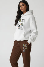 Woman wearing a white hoodie with colorful text design, holding a coffee cup against a plain background