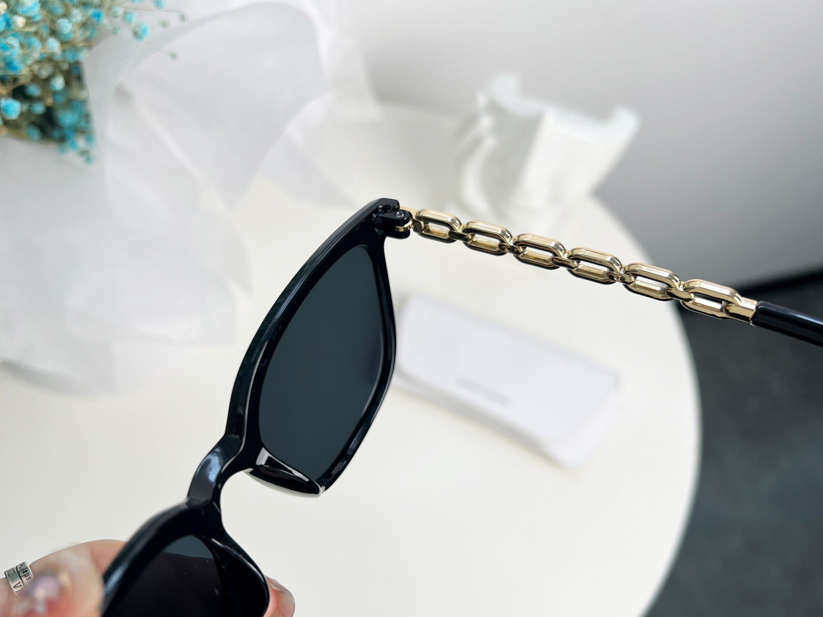 Black sunglasses with a gold chain held by a hand against a blurred background
