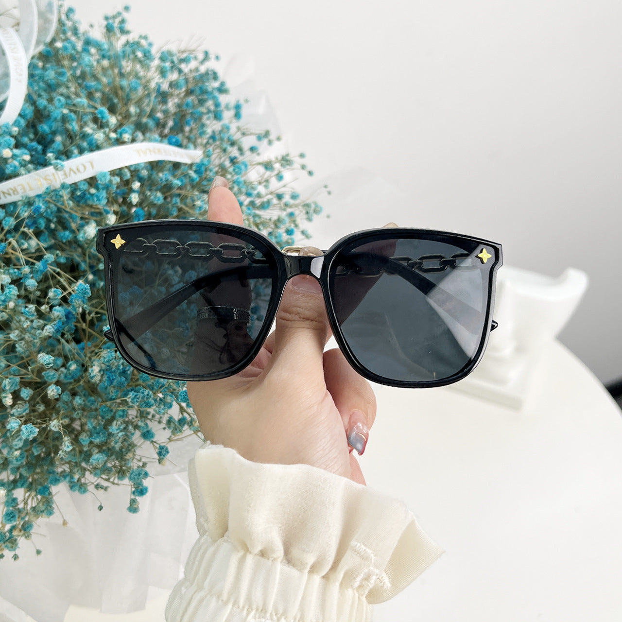 Black sunglasses held by a hand with a blurred background of blue flowers and a white surface.