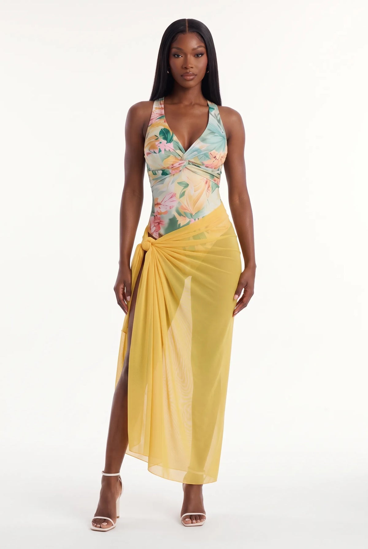 A model wearing a sleeveless wrap-style maxi dress with a pastel floral print bodice and a solid yellow draped skirt, paired with clear high-heeled sandals, against a plain white studio background.
