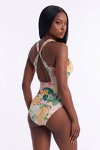 A woman modeling a one-piece swimsuit from the back, featuring a colorful floral print in shades of green, pink, yellow, and beige on a white studio background.