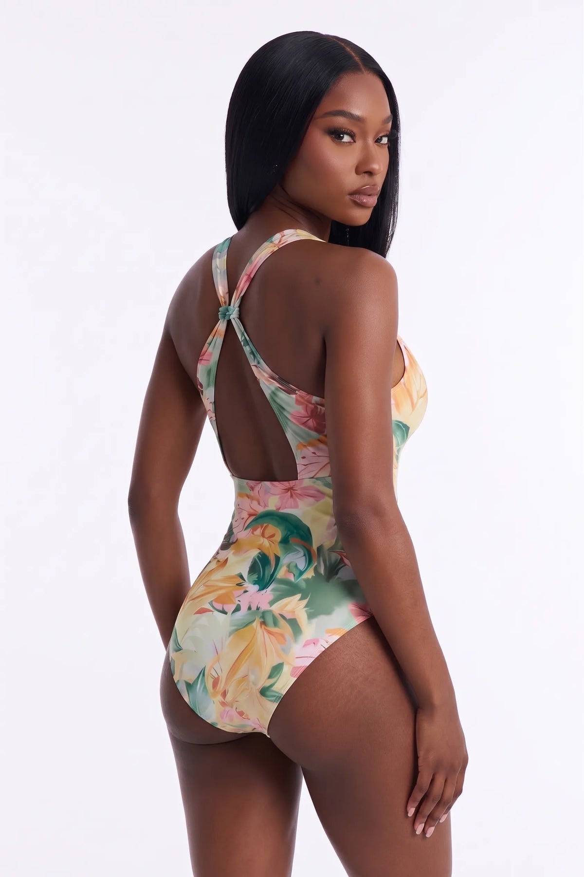 A woman modeling a one-piece swimsuit from the back, featuring a colorful floral print in shades of green, pink, yellow, and beige on a white studio background.
