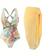A women's two-piece swimwear set featuring a tropical floral print one-piece swimsuit with crossover straps and a matching sheer yellow wrap sarong, displayed on a clean white background.
