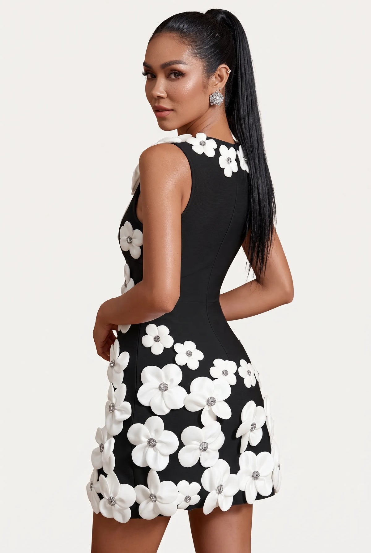A woman modeling a black bodycon mini dress with large white 3D floral appliqués on the bodice and skirt, paired with white heeled sandals, against a plain white studio background.