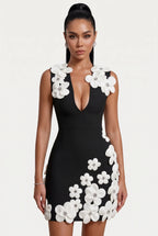 A woman modeling a black bodycon mini dress with large white 3D floral appliqués on the bodice and skirt, paired with white heeled sandals, against a plain white studio background.