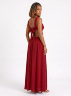 The dress shown is a crimson red maxi dress featuring side cutouts at the waist, a structured sweetheart bustier top with gathered drape detail, wide shoulder straps, and a bow tie backclosure.