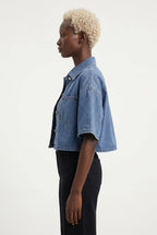 This is a medium-wash denim short-sleeved cropped jacket features a boxy, modern silhouette with relaxed drop shoulders and wide, elbow-length sleeves with stitched turned hems.