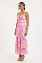 This is a vibrant maxi dress features an all-over abstract watercolor floral print in shades of pink, orange, purple, and yellow on a light background. It is designed with a sweetheart bustier-style top with sweetheart cutout at the center bust, thin shoulder straps, and a front waist cutout.