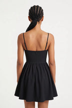 A woman viewed from the back wearing a black spaghetti-strap mini dress with a fitted bodice and flared skirt, against a plain light background.