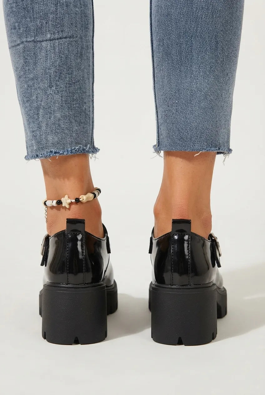Black patent leather Mary Jane platform shoes with buckle straps, shown from the back on a model wearing blue jeans and a beaded ankle bracelet.