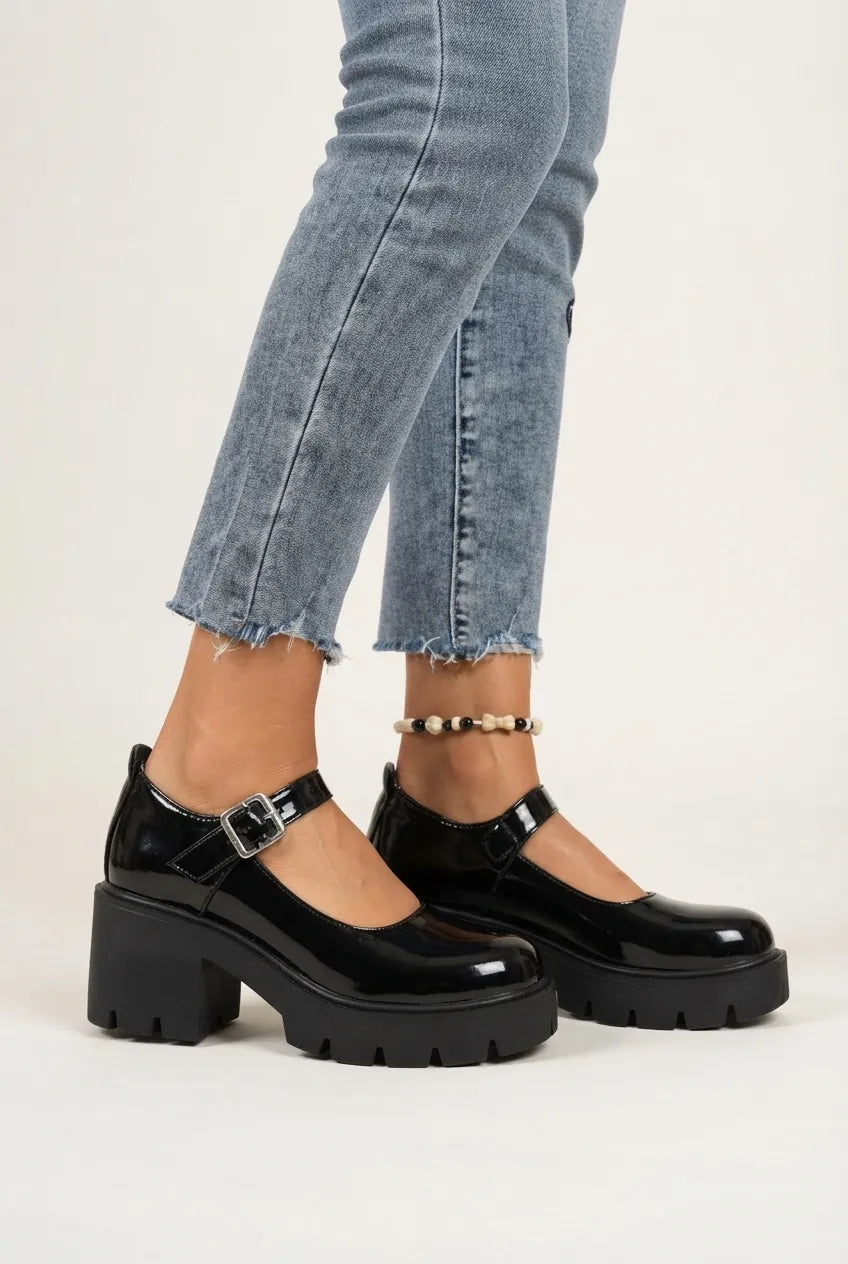 A person wearing black patent leather Mary Jane platform shoes with chunky lug soles and buckle straps, paired with light blue frayed-hem jeans against a plain light background.