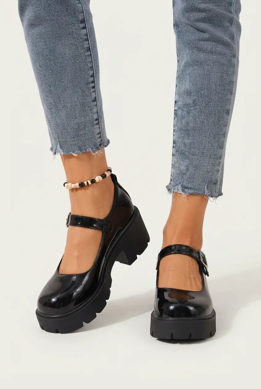 A person wearing black patent leather Mary Jane platform shoes with chunky lug soles, paired with light-wash frayed-hem jeans and a beaded ankle bracelet, shown against a plain white studio background.