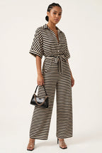 A woman modeling a black and white horizontally striped short-sleeve jumpsuit with a tie waist, holding a small black handbag, against a plain off-white studio background.
