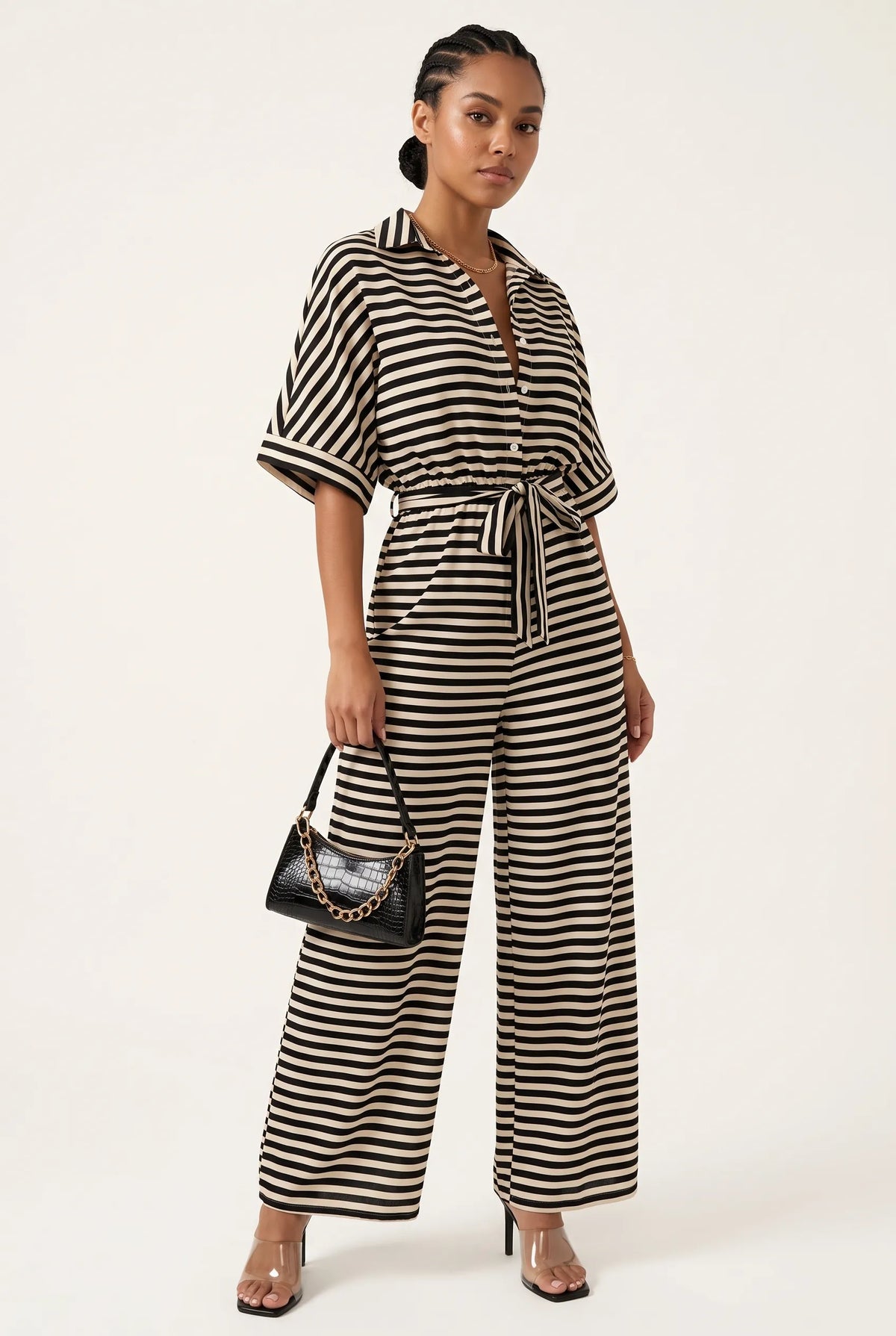 A woman modeling a black and white horizontally striped short-sleeve jumpsuit with a tie waist, holding a small black handbag, against a plain off-white studio background.