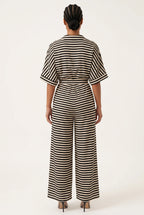 A woman viewed from the back wearing a black and beige horizontally striped short-sleeve jumpsuit with a matching waist tie, paired with black high heels, against a plain off-white studio background.