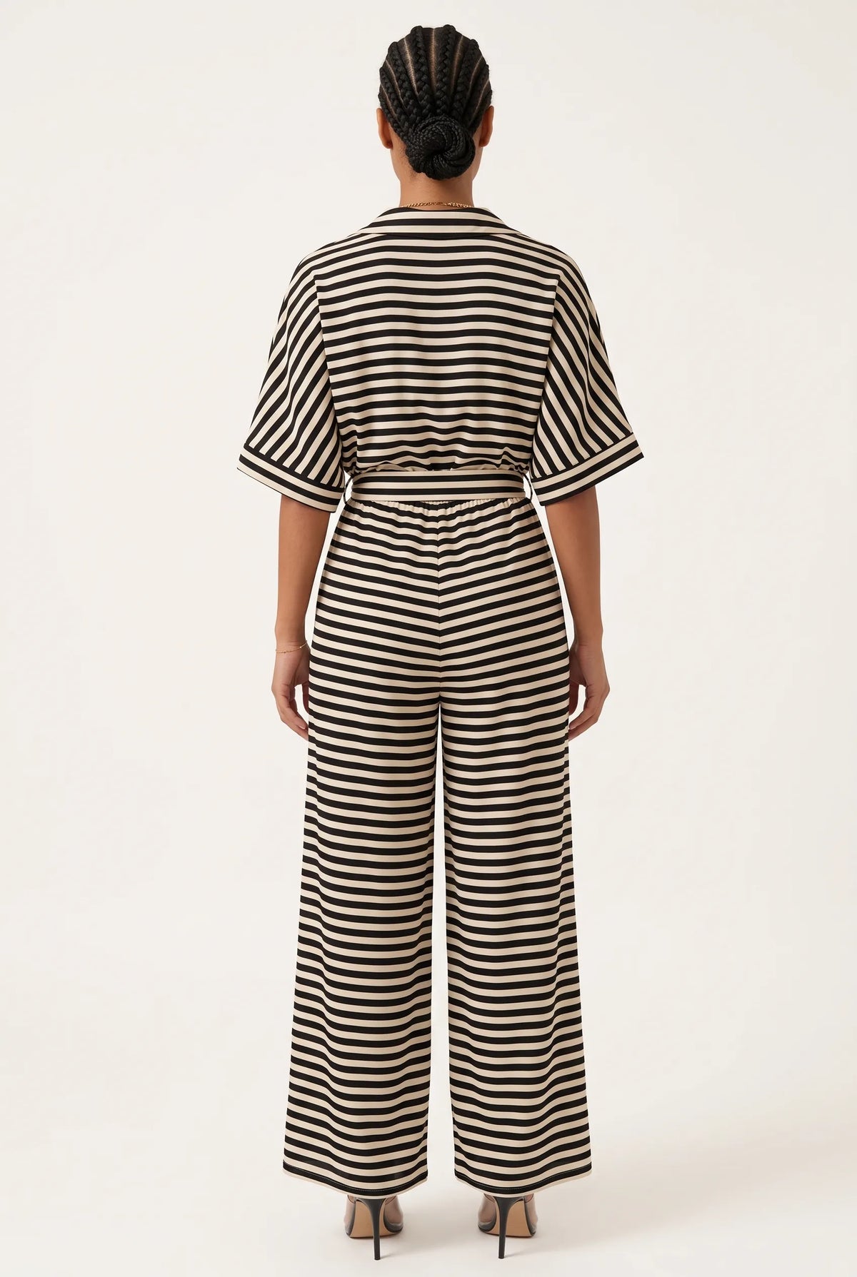 A woman viewed from the back wearing a black and beige horizontally striped short-sleeve jumpsuit with a matching waist tie, paired with black high heels, against a plain off-white studio background.