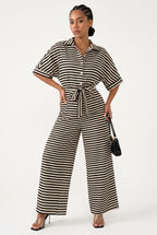 A woman modeling a black and white horizontally striped jumpsuit with short sleeves, a button-front collar, a tie waist, and wide legs, standing against a plain light background and holding a small black handbag.