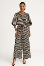 A woman modeling a black and white horizontally striped jumpsuit with a tie waist and wide legs, standing against a plain white studio background.