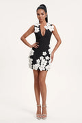 A woman modeling a black bodycon mini dress with large white 3D floral appliqués on the bodice and skirt, paired with white heeled sandals, against a plain white studio background.