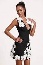 A woman modeling a black bodycon mini dress with large white 3D floral appliqués on the bodice and skirt, paired with white heeled sandals, against a plain white studio background.