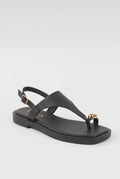 A black leather thong sandal with a low block heel, adjustable ankle strap with a gold buckle, and a small gold heart-shaped embellishment on the toe post, shown against a light gray background.