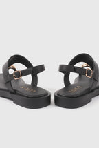 A pair of black platform sandals with double straps and gold-tone buckle details, shown from a side-rear angle on a light gray background.