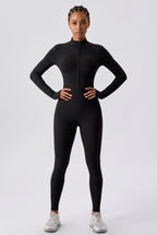 A model wearing a black long-sleeve full-zip athletic bodysuit or catsuit, paired with light gray sneakers, standing against a light gray studio background.