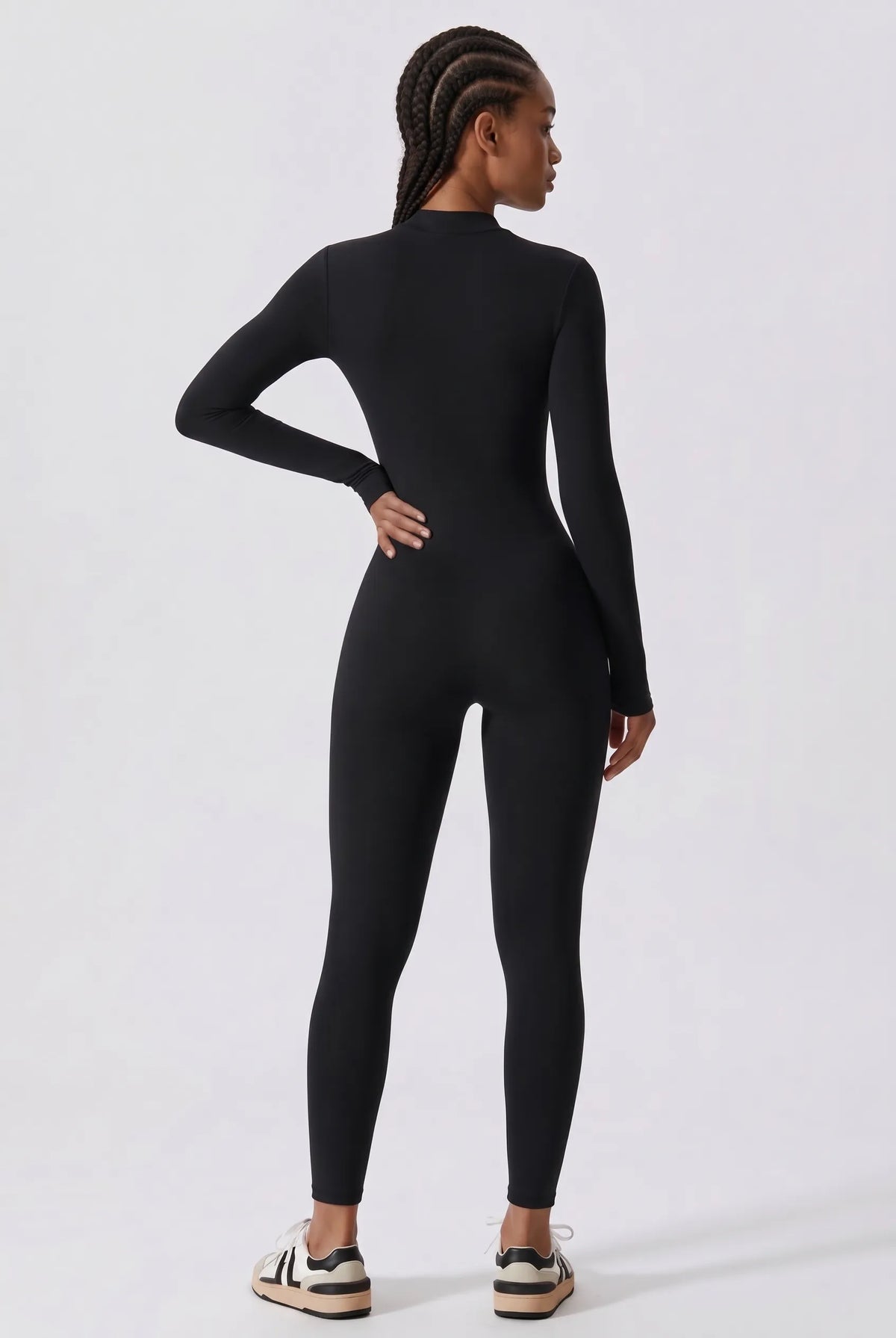 A woman modeling a black long-sleeve mock neck jumpsuit, shown from a rear three-quarter angle against a plain light gray studio background.