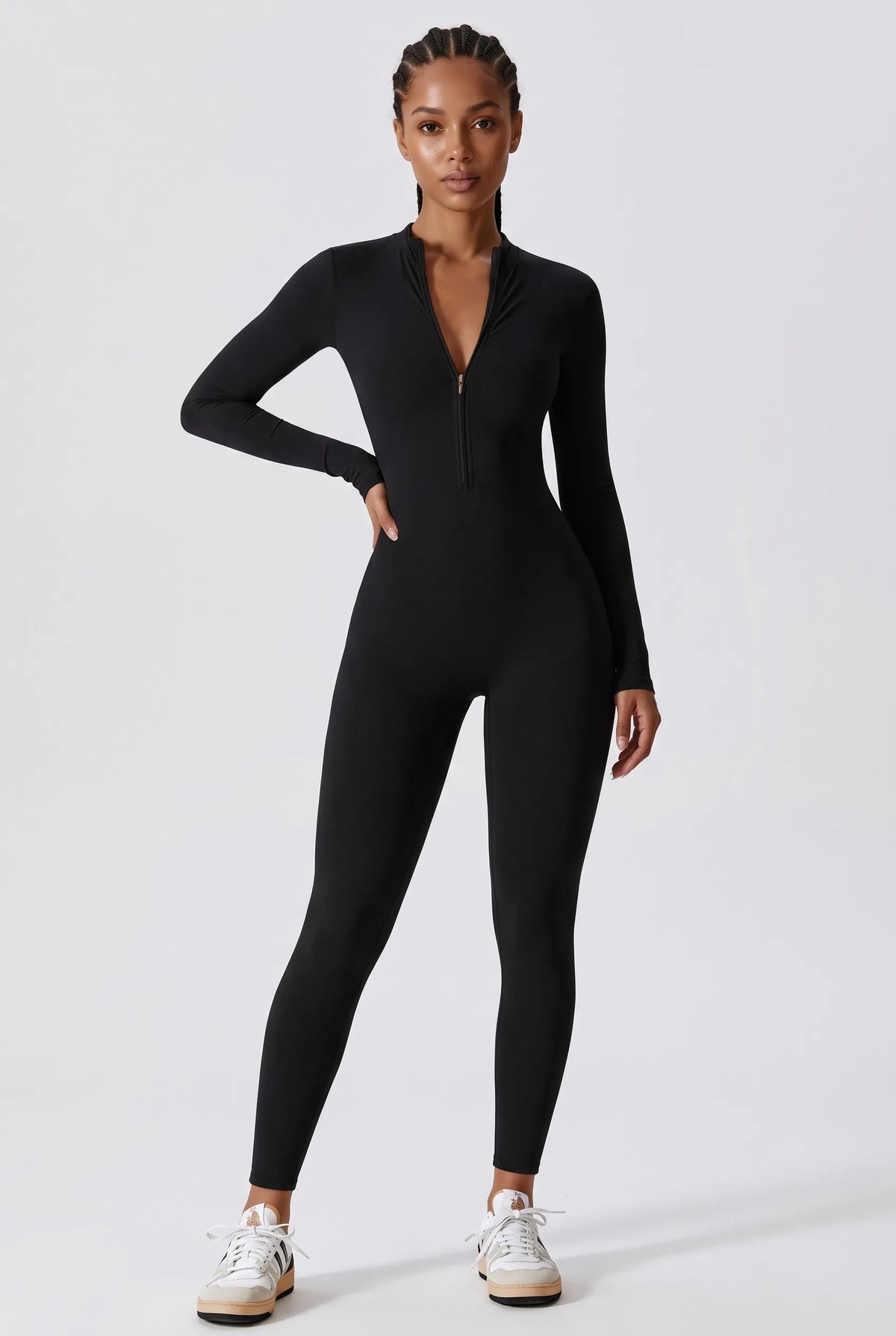 A model wearing a black long-sleeve zip-up bodysuit with a deep V-neckline, paired with white sneakers, standing against a light gray studio background.