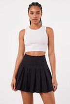 A woman modeling a white ribbed sleeveless crop top and a black pleated mini skirt against a plain light gray studio background.