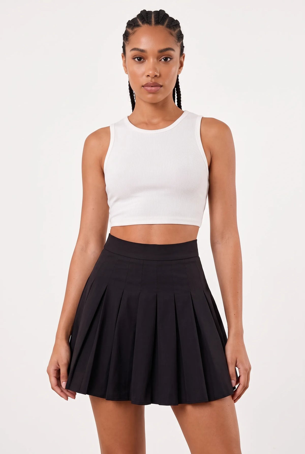 A woman modeling a white ribbed sleeveless crop top and a black pleated mini skirt against a plain light gray studio background.