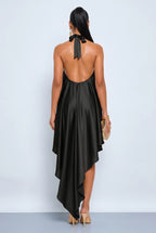 A woman viewed from the back wearing a black satin halter dress with an open back and tie detail at the neck, paired with beige heels and holding a small gold clutch against a light gray studio background.