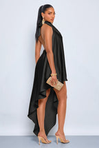 A woman modeling a black high-low dress with a draped asymmetrical neckline and open back, paired with nude high heels and a gold clutch, against a light gray studio background.