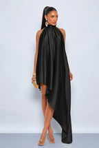 A model wearing a black satin halter-neck dress with an asymmetrical draped hem, styled with gold hoop earrings, a gold clutch, and clear high-heeled sandals against a light gray studio background.