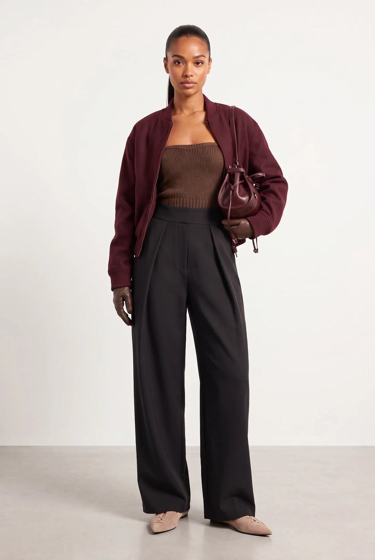 A model stands against a plain light gray studio background wearing a burgundy cropped jacket over a brown ribbed top, black wide-leg pleated trousers, and beige pointed flats, while carrying a matching burgundy handbag.