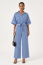 A woman modeling a blue and white horizontally striped short-sleeve tie-front jumpsuit with wide-leg cropped pants, holding a small black handbag against a plain light gray studio background.