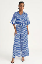 A woman modeling a blue and white horizontally striped short-sleeve jumpsuit with a matching tie belt, photographed against a plain light background.